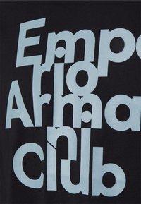 Černé tričko s velkým, světle modrým, překrývajícím se textem, který hlásá "Emporio Armani Club", s různou typografií a tvary.