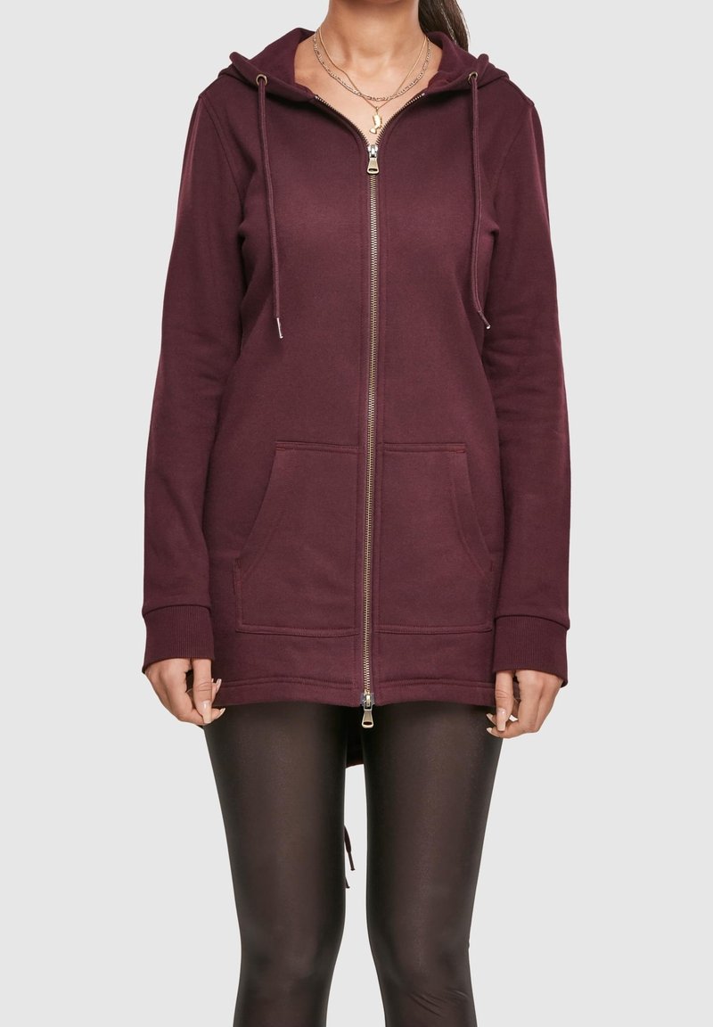 Femme portant un long sweat à capuche zippé bordeaux foncé avec poches avant et leggings noirs, debout sur un fond gris clair uni.