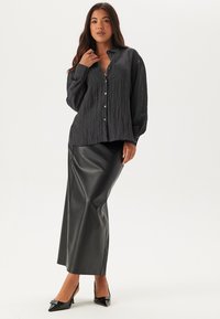 Camicia nera con bottoni, realizzata in tessuto texture, abbinata a una lunga e aderente gonna in pelle nera. La modella indossa tacchi alti neri.