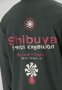 Pilka ilgomis rankovėmis marškinėliai su drąsiai spausdintu tekstu: "Shibuya dizaino paroda" ir spiralės bei saulės šviesos grafika rožinėje ir kreminėje spalvose.