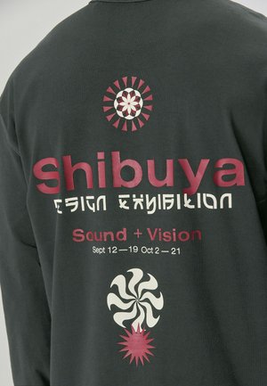 Grå langærmet skjorte med fed trykt tekst: "Shibuya Design Exhibition" og grafik af en spiral og solstråler i pink og creme.