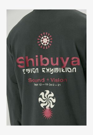 Gray long-sleeve shirt featuring bold printed text: "Shibuya Design Exhibition" and graphics of a spiral and sunburst in pink and cream.
