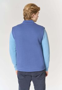 Gilet blu dal design senza maniche, con maniche lavorate a maglia di colore azzurro chiaro. Tessuto liscio con polsini a coste. Indossato sopra pantaloni scuri.
