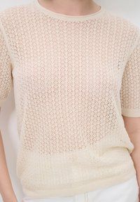 Helle beige gestrickte Pullover mit einem strukturierten Muster, kurzen Ärmeln und einem runden Halsausschnitt. Der Stoff wirkt halbtransparent mit gerippten Kanten.