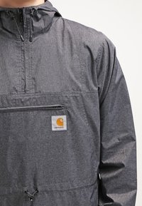 Szara, lekka kurtka z kapturem, z przednią kieszenią na zamek błyskawiczny oraz naszywką z logo Carhartt. Gładka tekstura, casualowy design i dół z ściągaczem.
