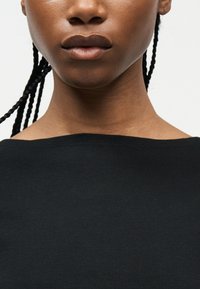 Κοντινό πλάνο του κάτω μέρους του προσώπου, του λαιμού και του άνω στήθους ενός ατόμου με πλεξούδες μαλλιά και μαύρη μπλούζα με λαιμόκοψη σε σχήμα βάρκας.