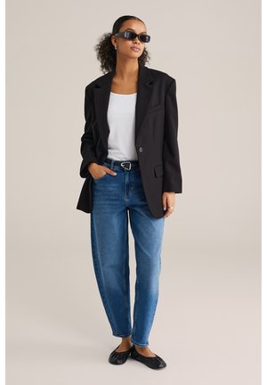 Blazer noir oversize sur un t-shirt blanc, jeans taille haute bleus et ballerines noires ; portant des lunettes de soleil sombres et des boucles d'oreilles en argent.