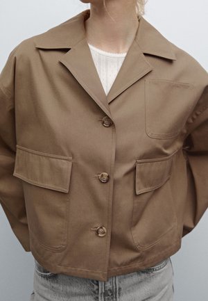 Frau trägt eine braune, bauchfreie Jacke mit großen Fronttaschen über einem weißen, gemusterten Oberteil und hellblauen Jeans.