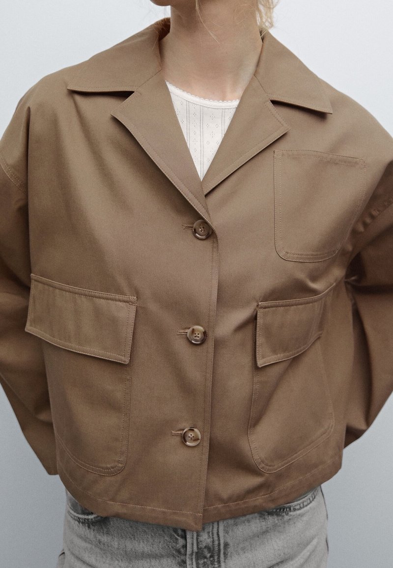 Femme portant une veste marron courte à boutons avec de grandes poches avant, par-dessus un haut blanc à motifs et un jean bleu clair.