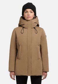 Parka marrone isolata con cappuccio, stringhe e tasche laterali con zip. Texture liscia e vestibilità standard. Berretto nero di maglia indossato.