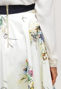Jupe midi florale en tissu blanc avec des imprimés botaniques colorés, dotée d'une taille dark et d'une fermeture éclair visible au dos.