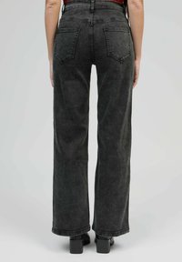 Personne portant un jean noir taille haute et large ainsi que des chaussures à talons bloc noires, se tenant de dos contre un fond clair.