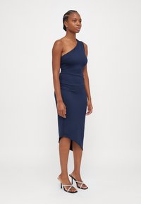 Robe asymétrique bleu marine, à une épaule, avec une coupe ajustée et un ourlet irrégulier. Tissu lisse avec un léger éclat ; associée à des talons à lanières.