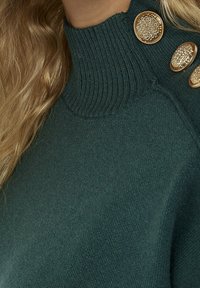 Mörkgrön tröja med hög ribbad krage och tre dekorativa guldknappar på axeln. Mjuk, slät textur med en figurnära siluett.