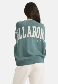 Sudadera verde de gran tamaño con "BILLABONG" impreso en grandes letras blancas en la parte posterior, con un escote redondo y puños acanalados.