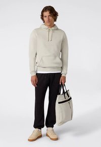 Felpa con cappuccio beige chiaro con tasca a marsupio e coulisse nere; abbinata a pantaloni della tuta neri e una borsa a spalla grigio chiaro con dettagli neri.