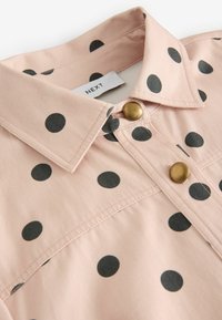 Camisa de manga corta rosa con lunares negros, que presenta un cuello estructurado y botones de latón, hecha de una tela suave y ligera.
