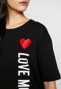 Svart t-shirt med korta ärmar, med en röd hjärt-grafik och fet vit text som säger "ÄLSKA MIG" tryckt vertikalt på sidan.