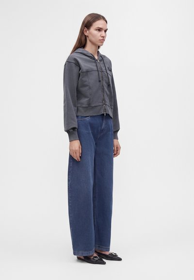 Jeune femme debout de profil, portant un sweat à capuche gris zippé, un jean large bleu et des ballerines noires pointues sur fond uni.