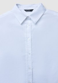 Weiße Button-down-Hemden mit spitzem Kragen, strukturiertem Stoff, sechs Knöpfen auf der Vorderseite und einem Markenlabel am Kragen.