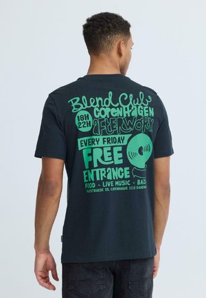 Man in zwart T-shirt met groene tekst die een gratis vrijdag na het werk evenement in Kopenhagen promoot met eten, live muziek en bazaar van 18.00 tot 22.00 uur.