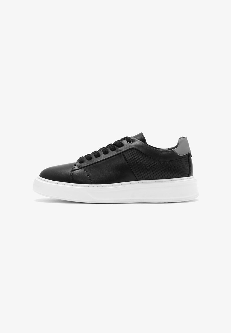 Zapatilla deportiva de cuero negro con un acabado suave, suela de goma blanca, puntera redonda y cordones negros. El panel lateral presenta un diseño texturizado.