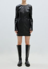 Zwart leren mini-jurk met lange mouwen, ronde halslijn en naden; gecombineerd met knie-hoge zwarte laarzen met een chunky profiel.