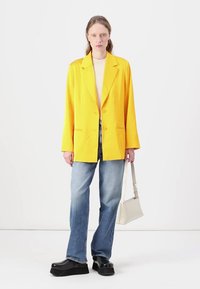 Gul blazer med spiss krage, to knapper og sidelommer, kombinert med lyseblå rette jeans og svarte platåklosser.