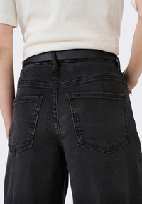 Person, der schwarze Denim-Jeans mit großen Gesäßtaschen und ein weißes, eingestecktes Hemd trägt, von der Taille abwärts, Rückansicht.