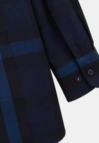 Chemise à carreaux bleu marine à manches longues, avec des poignets contrastants, deux boutons sur la manche et une texture en coton lisse.