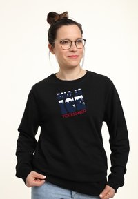 Schwarzer Sweatshirt mit einem grafischen Design, auf dem der Text "COLD AS ICE" in Blau und "FOREIGNER" in Rot zu sehen ist, vor einem strukturierten Hintergrund.