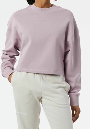 Persona que lleva una sudadera corta lila clara y pantalones deportivos color crema con el logo "new balance" en el muslo izquierdo, manos en los bolsillos.
