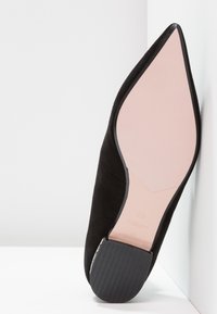 Zapato de ante negro con puntera afilada, suela rosa claro y tacón de goma negro. Presenta una textura suave y líneas limpias.