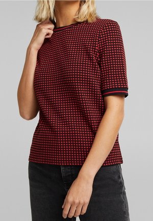 Mujer con una blusa de manga corta a cuadros roja y negra y vaqueros oscuros, con la mano cerca del cuello y cabello rubio visible.