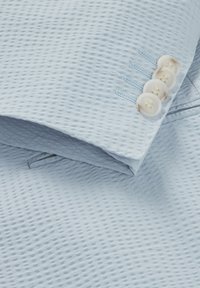 Ljusblå texturerad tygjacka ärm med fyra krämfärgade knappar och subtila sömdetaljer. Slät yta med ett rutmönster.