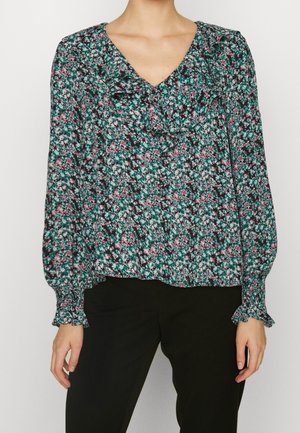 Femme portant un chemisier floral à manches longues avec col en V et détails à volants, associé à un pantalon noir, debout devant un fond uni.