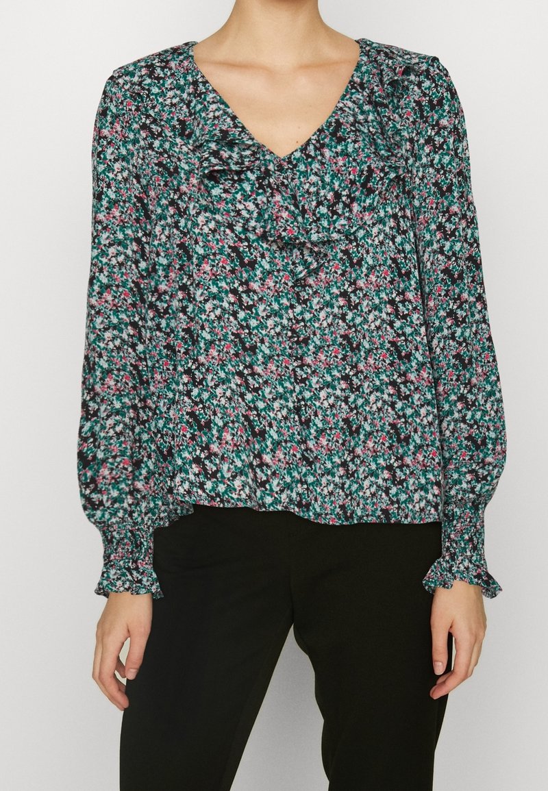 Femme portant un chemisier floral à manches longues avec col en V et détails à volants, associé à un pantalon noir, debout devant un fond uni.