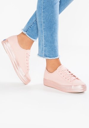 Persona che indossa jeans azzurri con orli sfrangiati e sneaker platform rosa chiaro, con un piede sollevato su sfondo bianco.