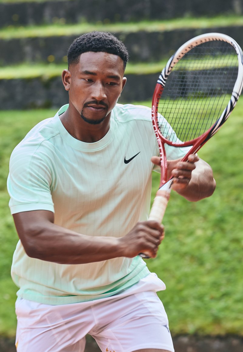 Tennisemängija helesinises, ribakujulises lühikestes varrukates särgis ja valgetes lühikeses, hoiab käes punast reketti, valmistudes palli löömiseks.