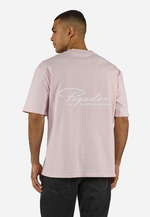 Mann trägt ein hellrosa übergroßes T-Shirt mit dem Schriftzug "Pegador All Rights Reserved" auf der Rückseite, kombiniert mit schwarzen Jeans, steht seitlich.