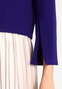 Fialové sako na míru s krátkým střihem a strukturovaným designem, doplněné jemnou plisovanou sukní v světle béžové barvě.