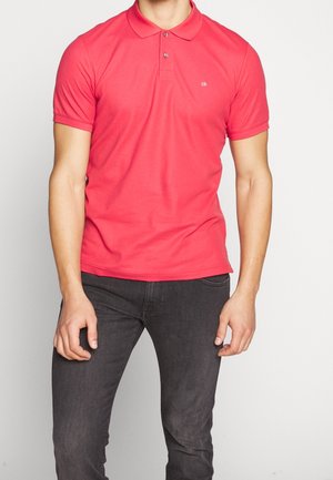 Rosa Poloshirt mit kurzen Ärmeln, aus glattem Baumwollmix, mit einer Knopfleiste mit zwei Knöpfen und einem kleinen Logo auf der Brust. Kombiniert mit dunklen Jeans.