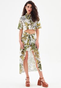 Completo da donna in due pezzi composto da un top corto floreale e una gonna maxi trasparente. I colori includono verde, giallo e bianco. Il materiale sembra leggero.