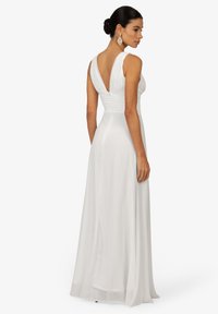 Robe blanche longue avec un corsage ajusté, des détails plissés et un dos en V profond. Texture lisse, larges bretelles et fermeture éclair dissimulée.