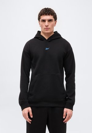 Młody mężczyzna w czarnej bluzie z kapturem z przednią kieszenią i małym niebieskim logo Reebok, stojący na jasnym, jednolitym tle.
