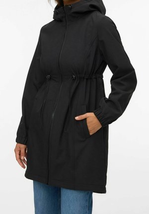 Kvinde iført lang sort hættejakke med lynlås foran, elastiske manchetter og snoretræk i taljen, hænderne i lommerne, kombineret med blå jeans.