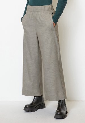 Beige wijde geruite broek met elastische tailleband, gedragen met zwarte stevige laarzen en een donkergroen top met lange mouwen, handen in de zakken.