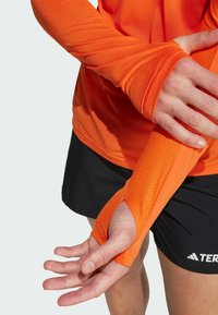 Langarm sportshirt van feloranje stof met een gestructureerd armontwerp en duimgatdetail, gecombineerd met een zwarte sportrok.