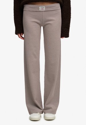 Persoon draagt bruine en beige dun gestreepte wijde broek met een Superdry-logo op de taille en witte sneakers.