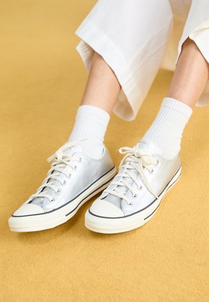 Baskets en toile argentées avec semelles en caoutchouc blanches, dotées de lacets et d'un bout arrondi. Tige texturée avec un léger éclat. Portées avec des chaussettes blanches à côtes.